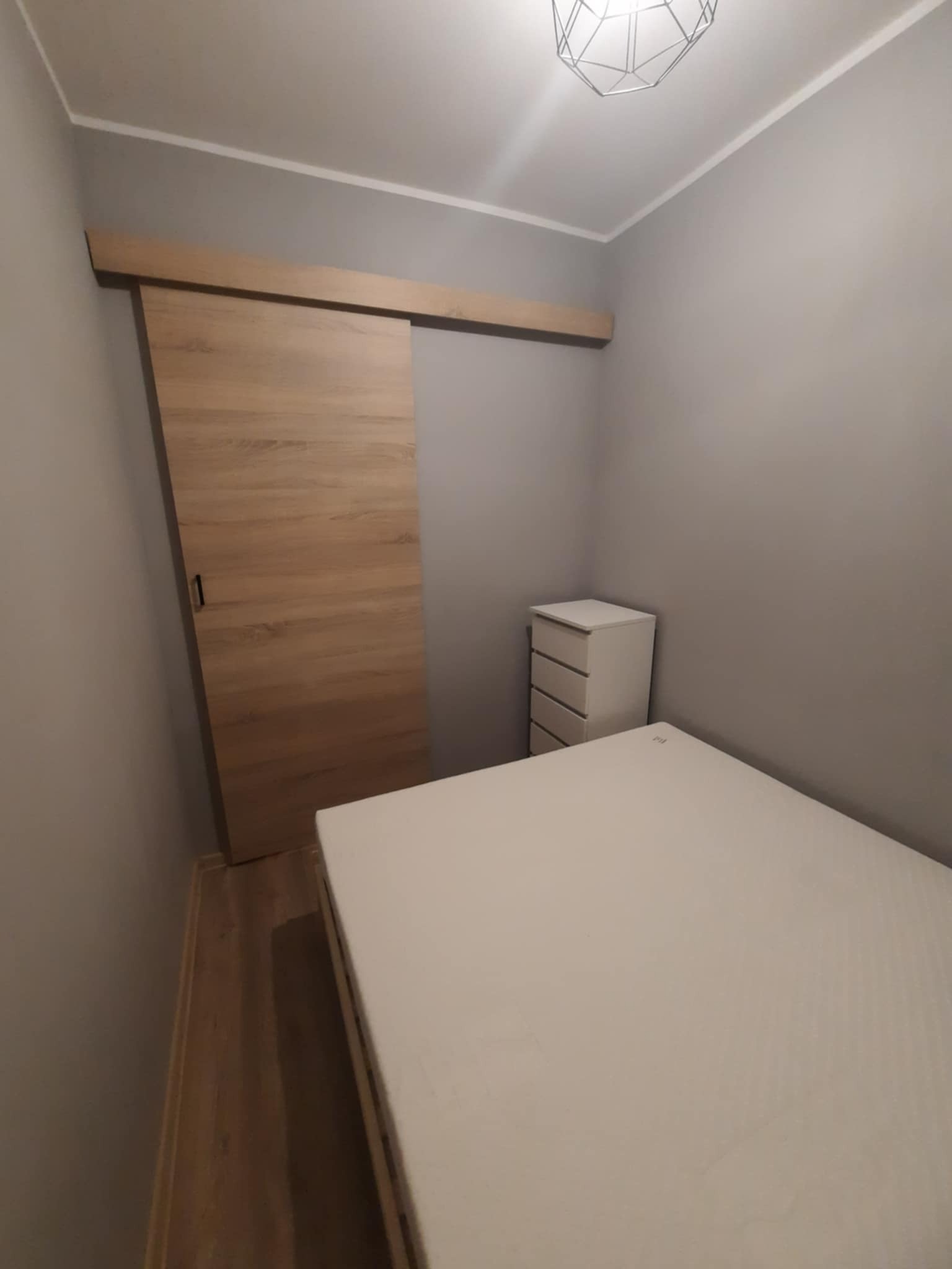 Zamknięte przesuwne drzwi z jasnego drewna, zamontowane na szynie, widoczne w sypialni z łóżkiem i białą komodą.