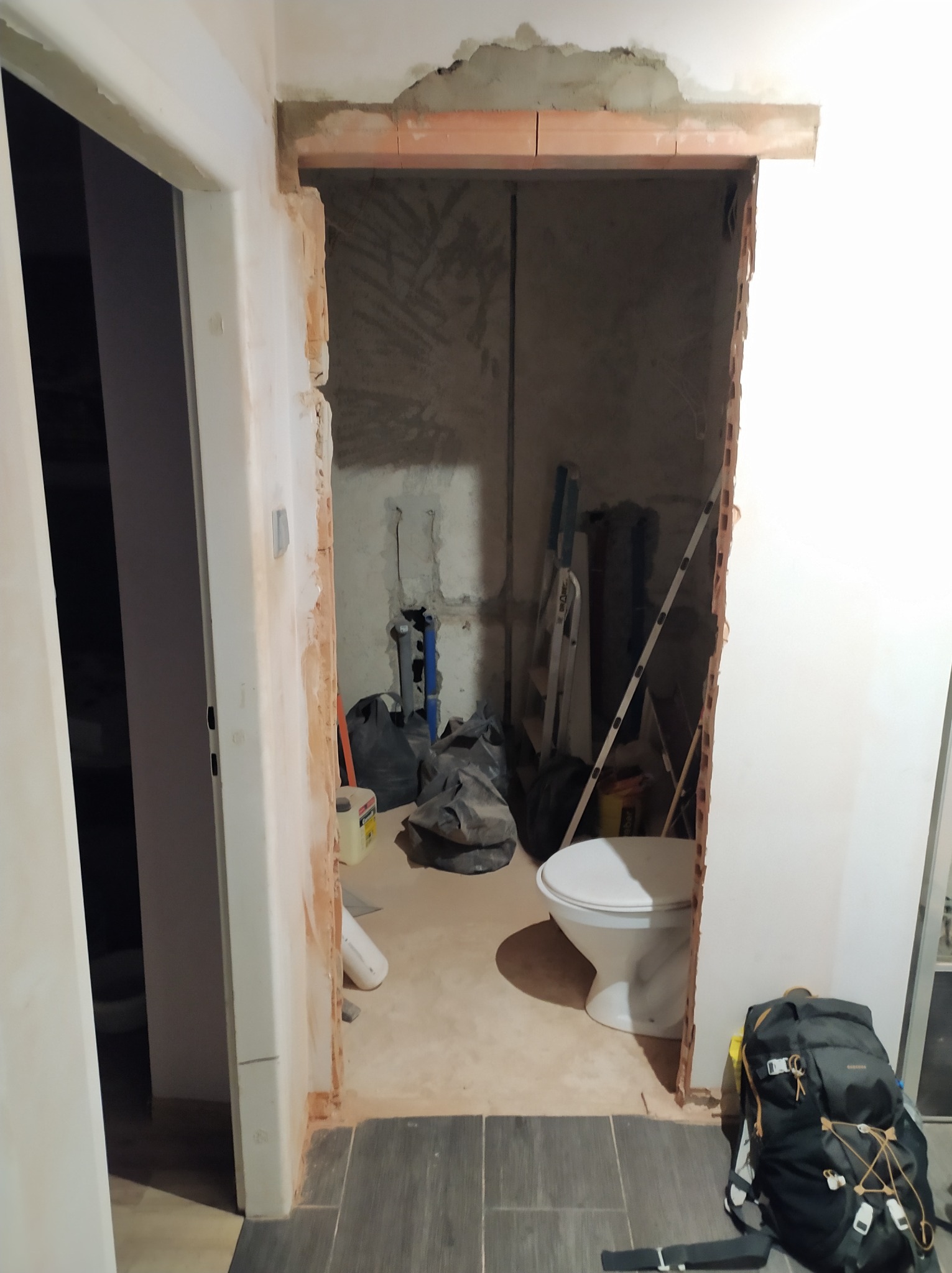 Remont łazienki w trakcie: widoczne instalacje, surowa ściana z cegieł nad otworem drzwiowym, częściowo ułożone płytki podłogowe, toaleta, drabina i torby z materiałami budowlanymi wewnątrz...