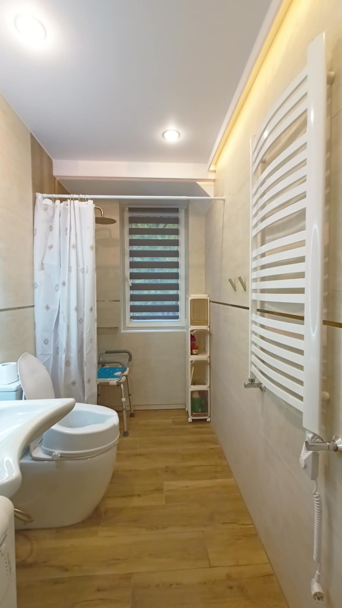 Jasna łazienka z beżowymi płytkami, drewnopodobną podłogą i białym grzejnikiem drabinkowym. Widoczny prysznic z zasłoną, okno z roletami i krzesło prysznicowe.