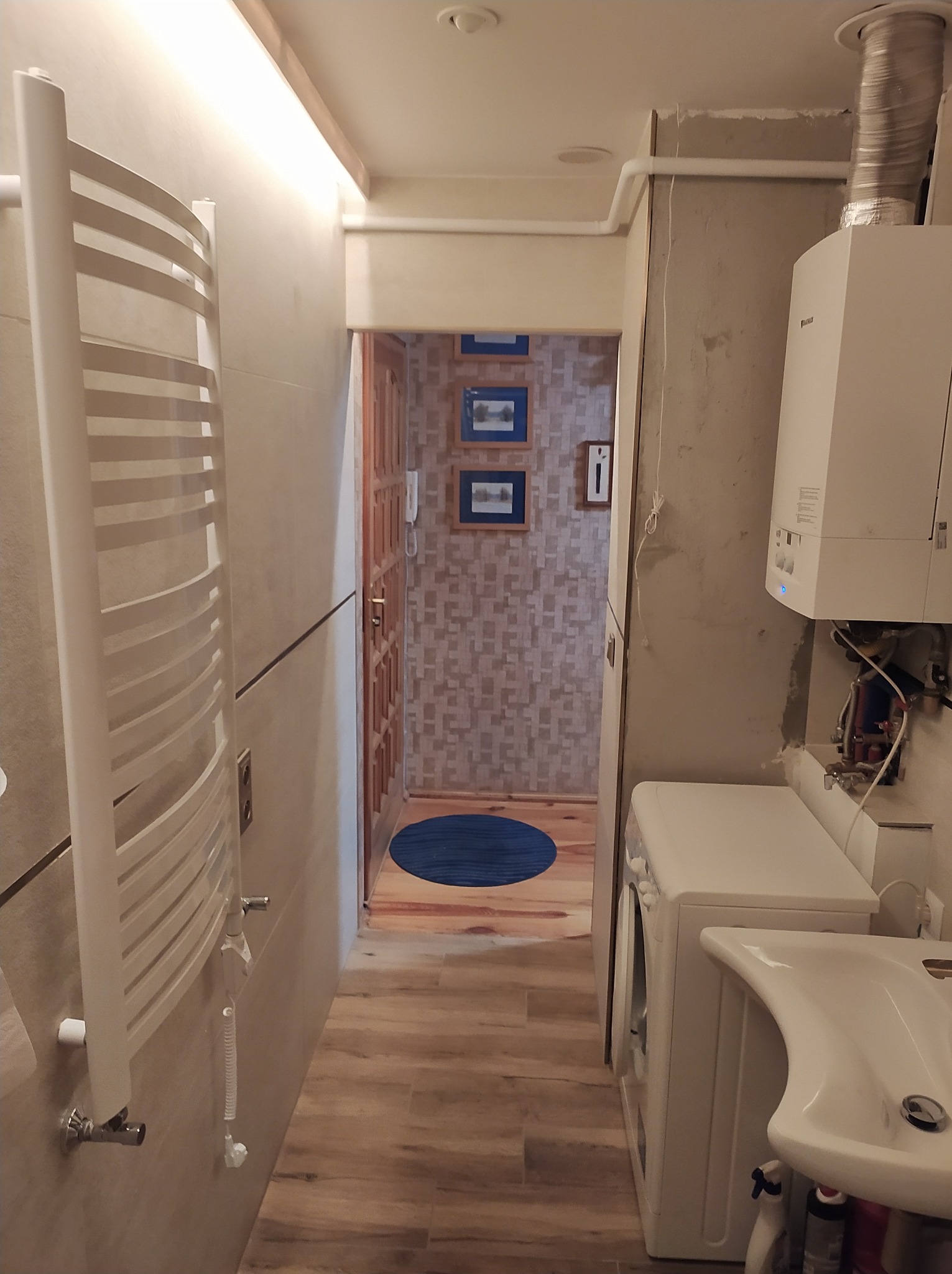 Wnętrze łazienki w trakcie remontu: grzejnik drabinkowy, pralka, umywalka i piec gazowy. Widoczne niedokończone ściany i instalacje. W tle korytarz z drewnianą podłogą.