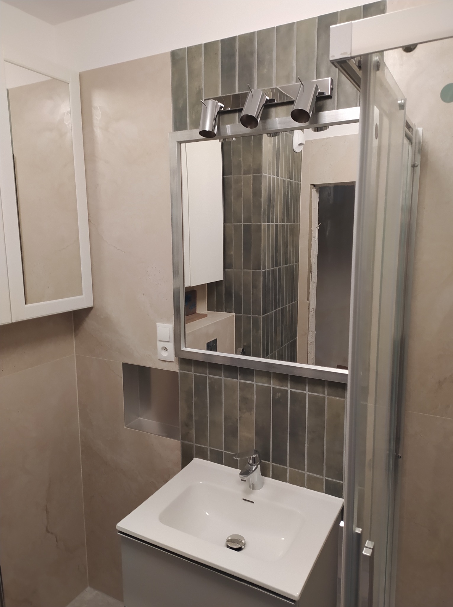 Nowoczesna łazienka po remoncie: biała umywalka z szafką, szklana kabina prysznicowa, lustro z oświetleniem i beżowo-szare płytki na ścianach. Widoczny fragment wnęki.