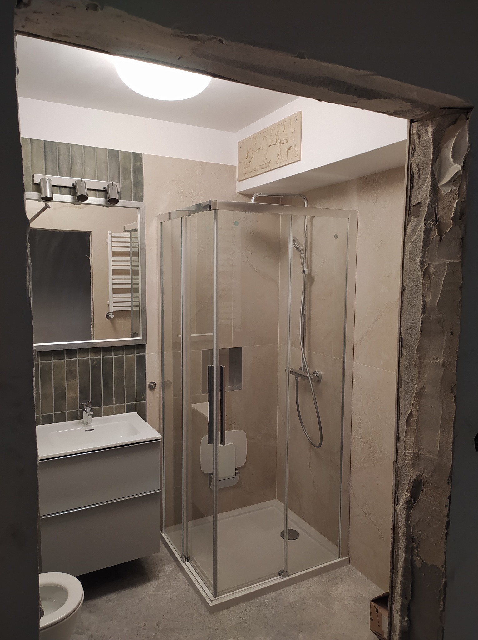 Wykończona łazienka z kabiną prysznicową, umywalką z szafką i toaletą. Widoczne beżowe płytki na ścianach i szare na podłodze. Fragment surowego otworu drzwiowego.