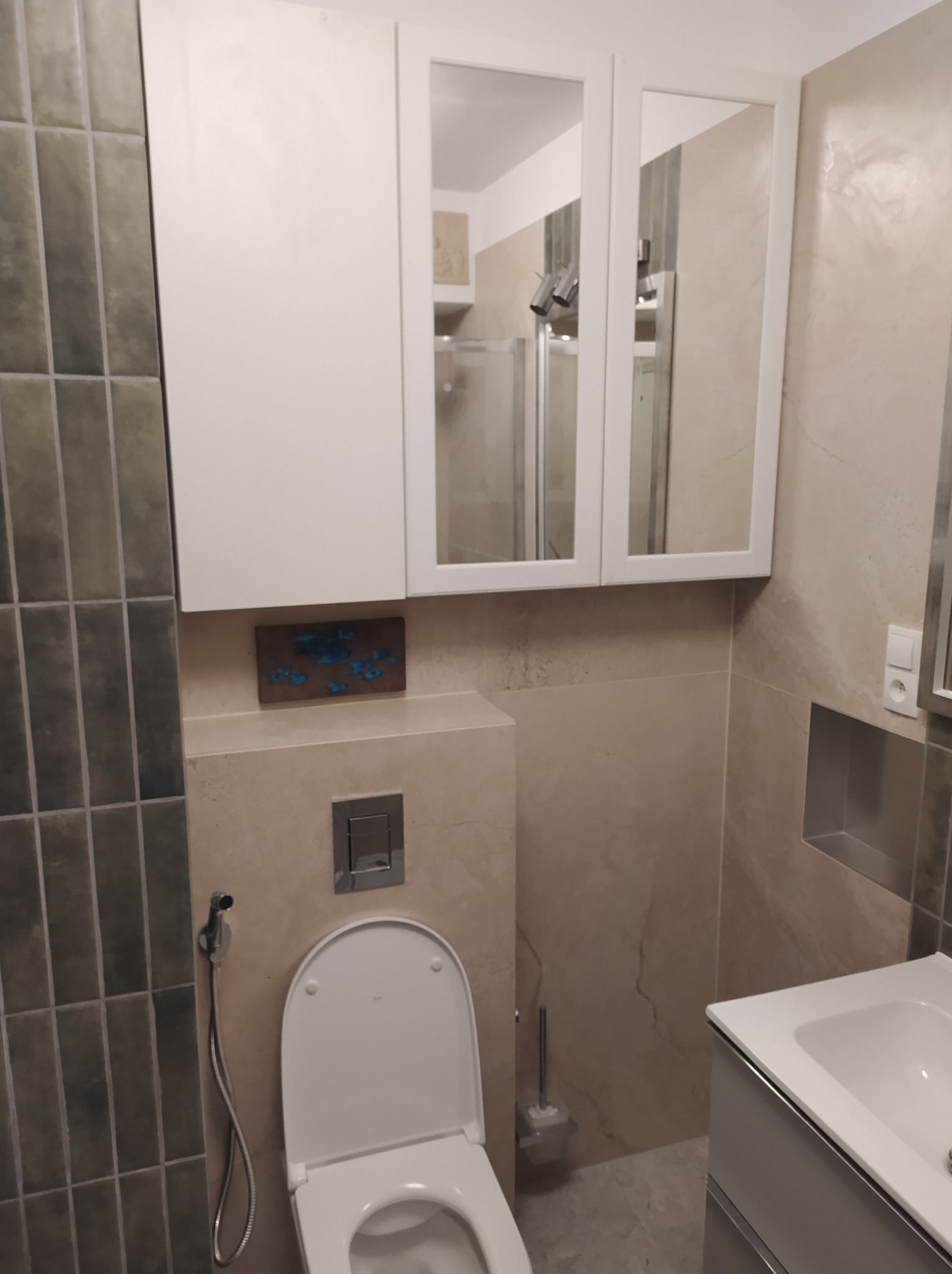 Nowoczesna łazienka z beżowymi płytkami, wiszącą szafką z lustrem, toaletą z bidetką i umywalką. Widoczna dekoracja ścienna z motywem niebieskich kwiatów.