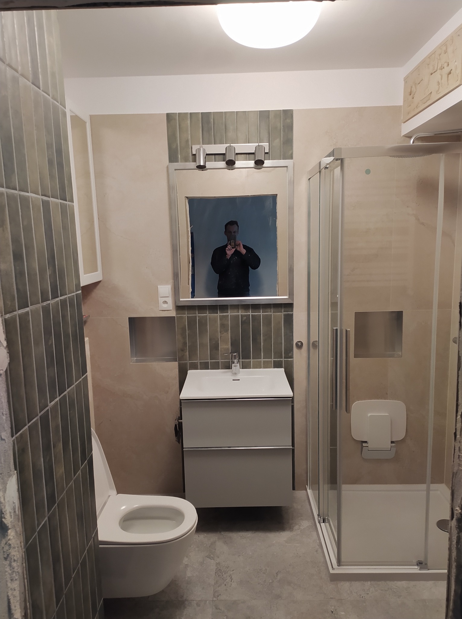 Wykończona łazienka z kabiną prysznicową, umywalką z szafką i toaletą. Widoczne szare płytki i lustro z odbiciem osoby robiącej zdjęcie.