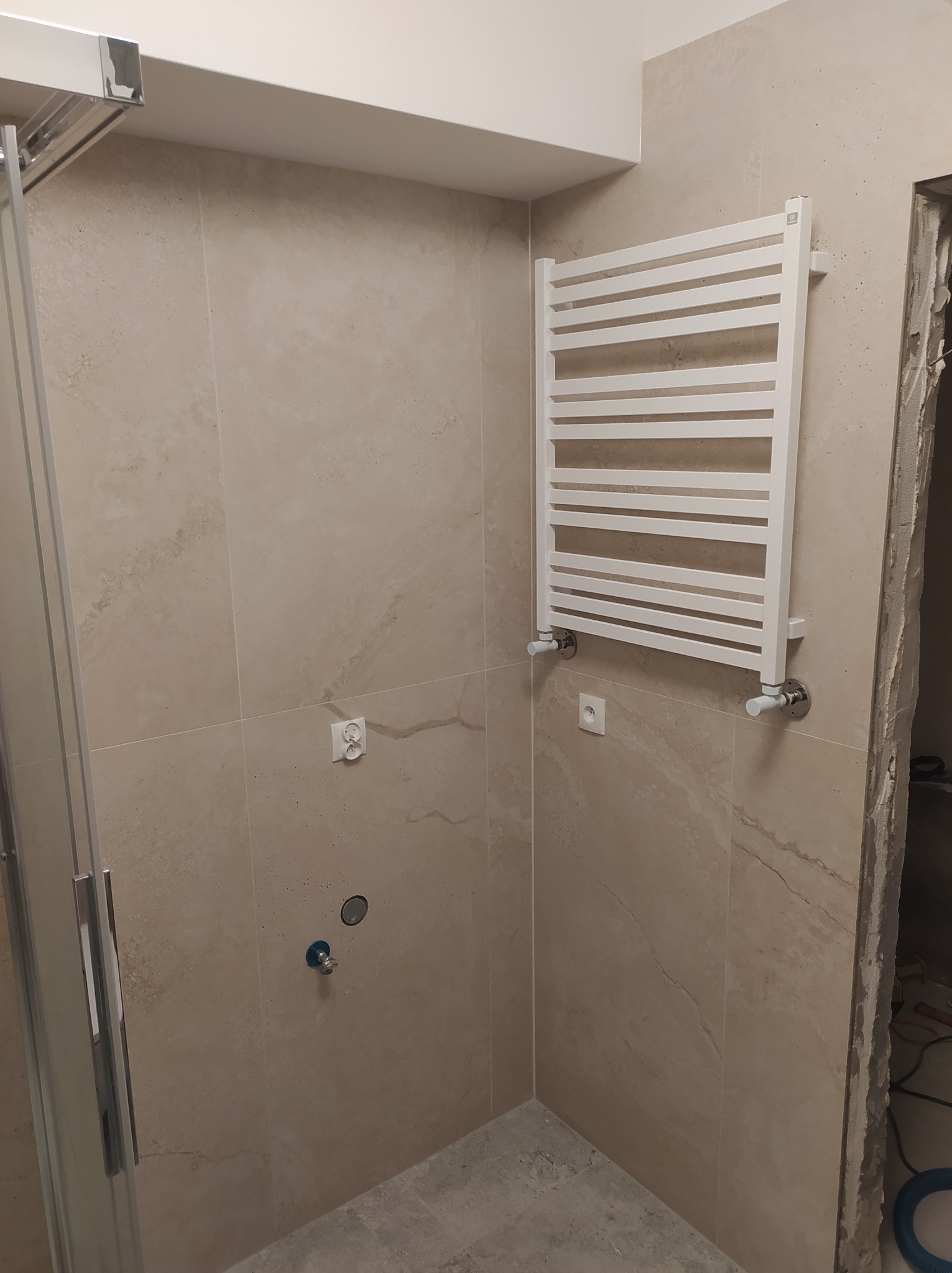 Beżowa łazienka w trakcie wykańczania: zamontowany grzejnik drabinkowy, widoczne przyłącza, fragment kabiny prysznicowej i niedokończone obramowanie drzwi.