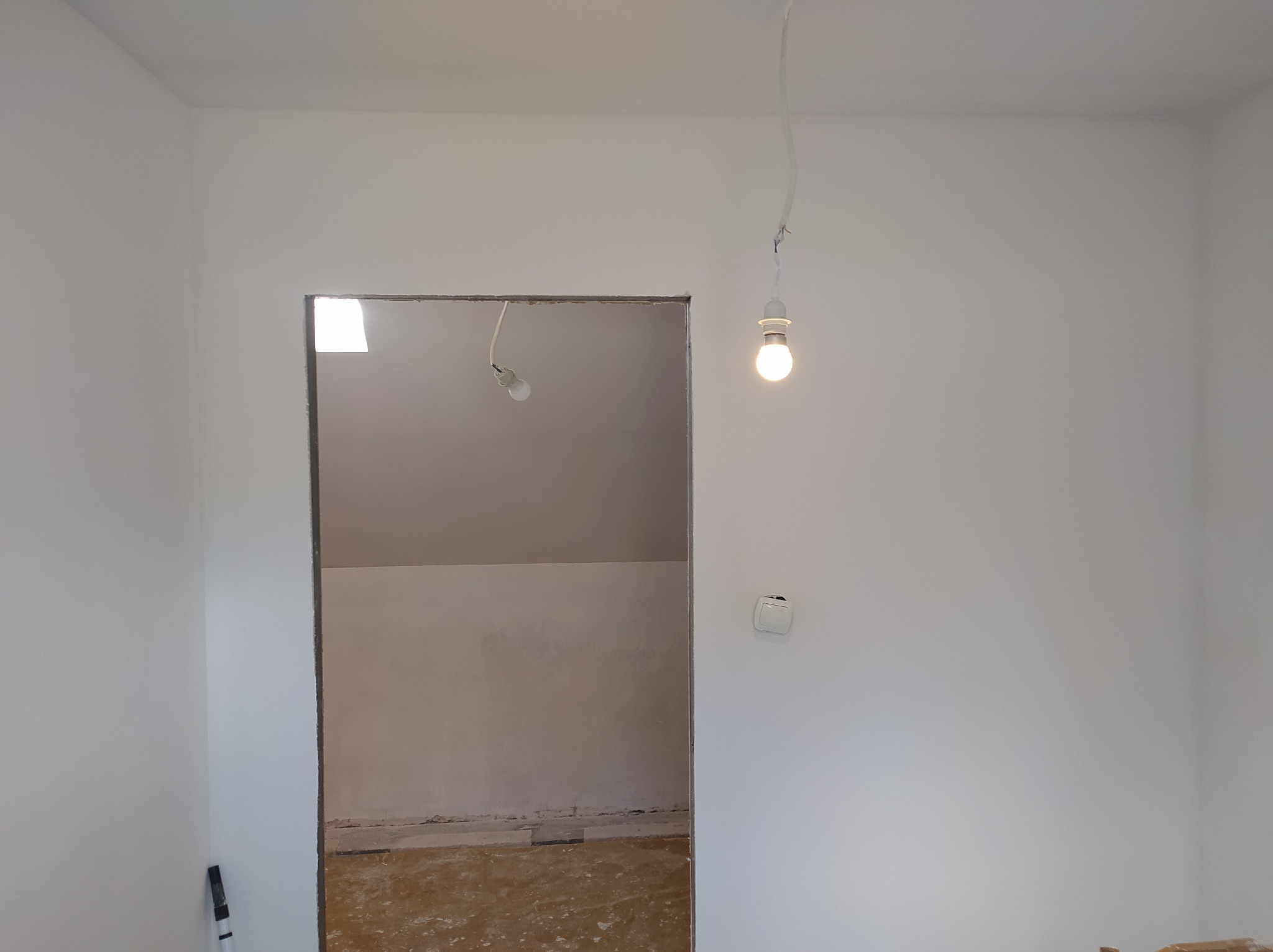 Białe ściany po szpachlowaniu z widocznym otworem drzwiowym i wiszącą żarówką. Wnętrze w trakcie wykańczania, surowy stan podłogi.