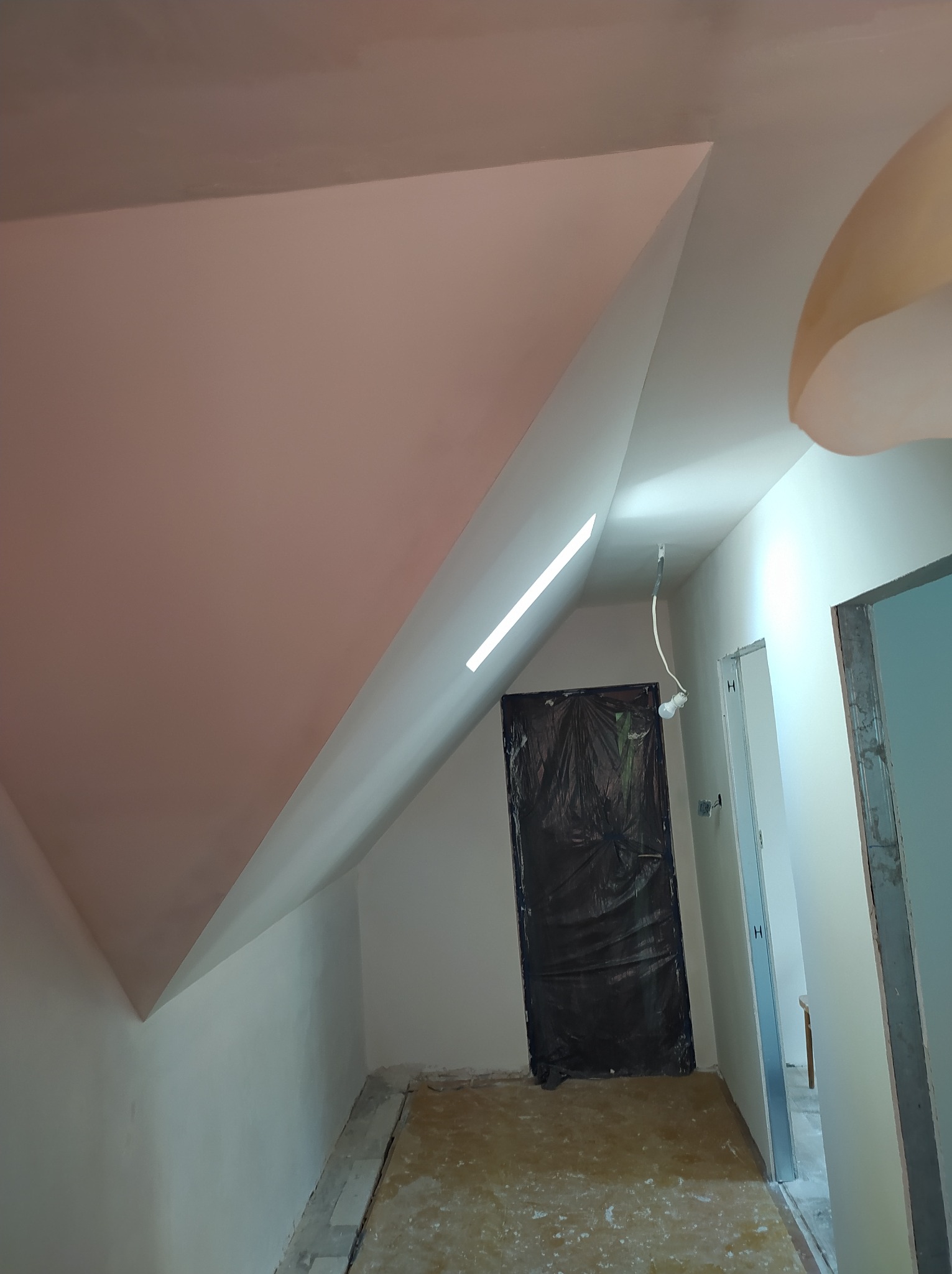 Wykończone poddasze ze skośnym sufitem, pomalowane na biało, z widocznymi drzwiami zabezpieczonymi folią i fragmentem oświetlenia. Widok z dołu.