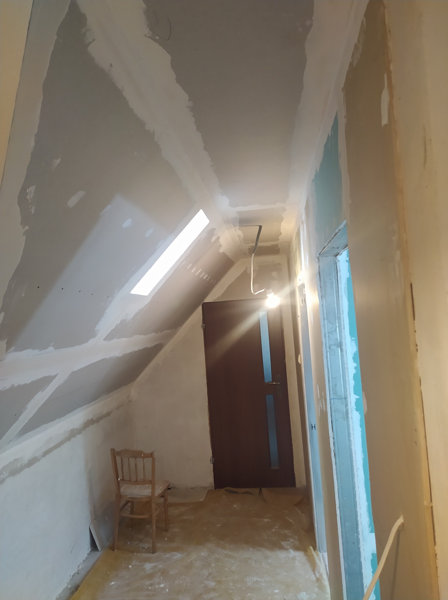 Poddasze w trakcie prac wykończeniowych: szpachlowane ściany, okno dachowe, widoczne płyty gipsowe, drewniane drzwi i krzesło. Wnętrze w stanie surowym, przygotowane do malowania.