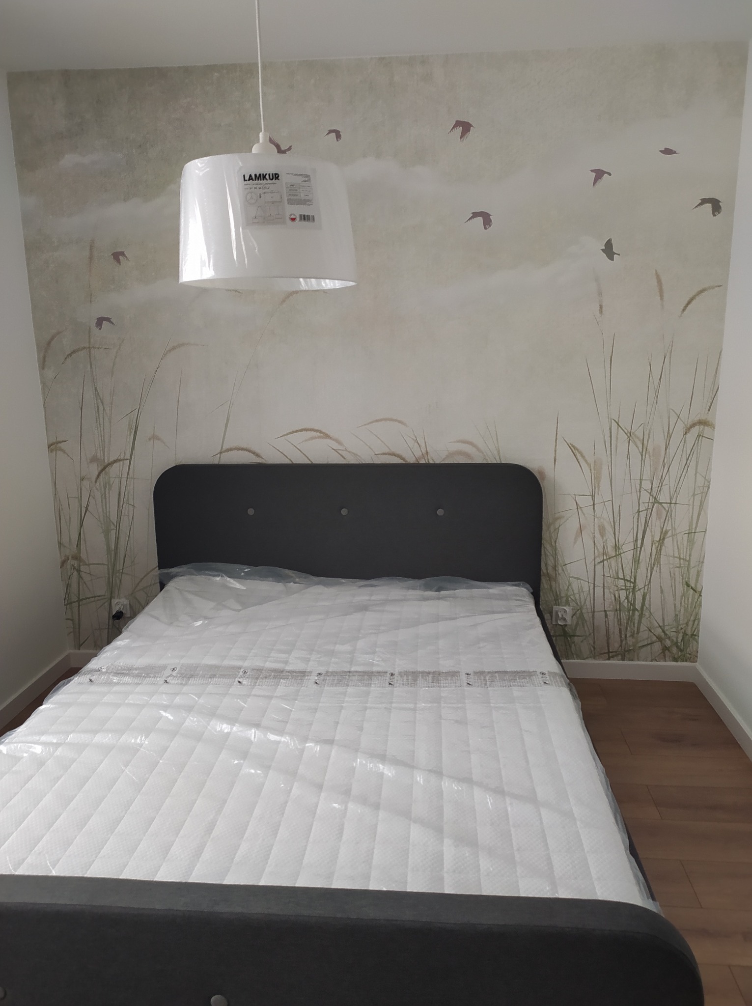 Szare łóżko z materacem w folii ochronnej, na tle ściany z motywem trawy i ptaków. Nad łóżkiem wisi biała lampa LAMKUR.