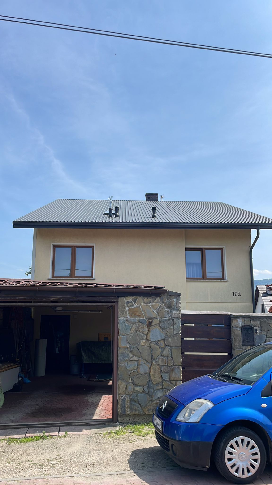 Dom jednorodzinny z szarym, metalowym dachem falistym i brązowymi oknami. Widoczny garaż z kamienną elewacją i fragment niebieskiego samochodu.