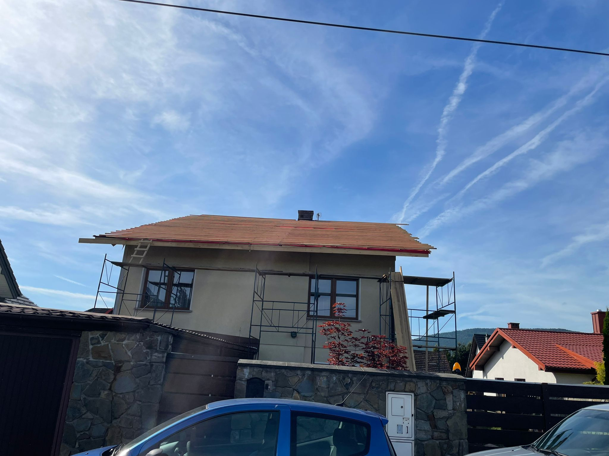 Dom w trakcie krycia dachu, widoczne rusztowania i częściowo ułożona dachówka. Błękitne niebo z smugami kondensacyjnymi w tle.