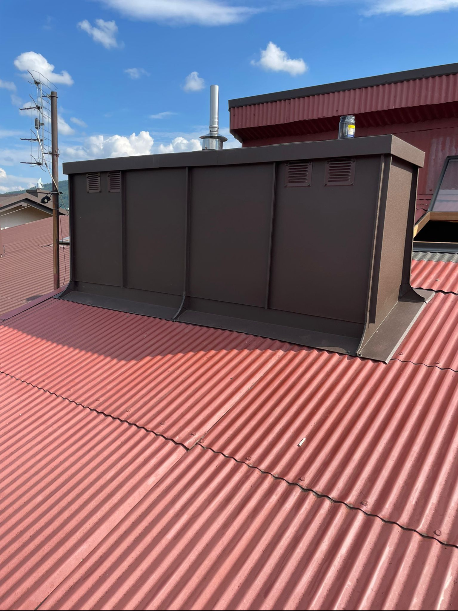 Czerwony dach z blachy falistej z brązową obudową wentylacyjną. Widoczne łączenia blach i elementy mocujące. W tle błękitne niebo z chmurami.