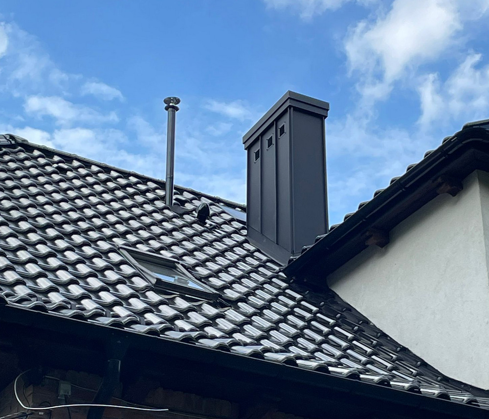 Czarny komin z blachy na dachu pokrytym ciemną dachówką ceramiczną, widok z dołu na tle błękitnego nieba z chmurami. Dach z oknem połaciowym.