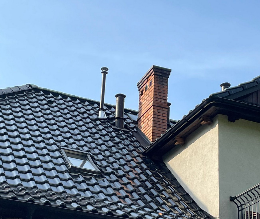 Błyszczący, ciemny dach z dachówki ceramicznej, widoczne kominy (jeden z cegły), okno dachowe i fragment elewacji budynku w słoneczny dzień.