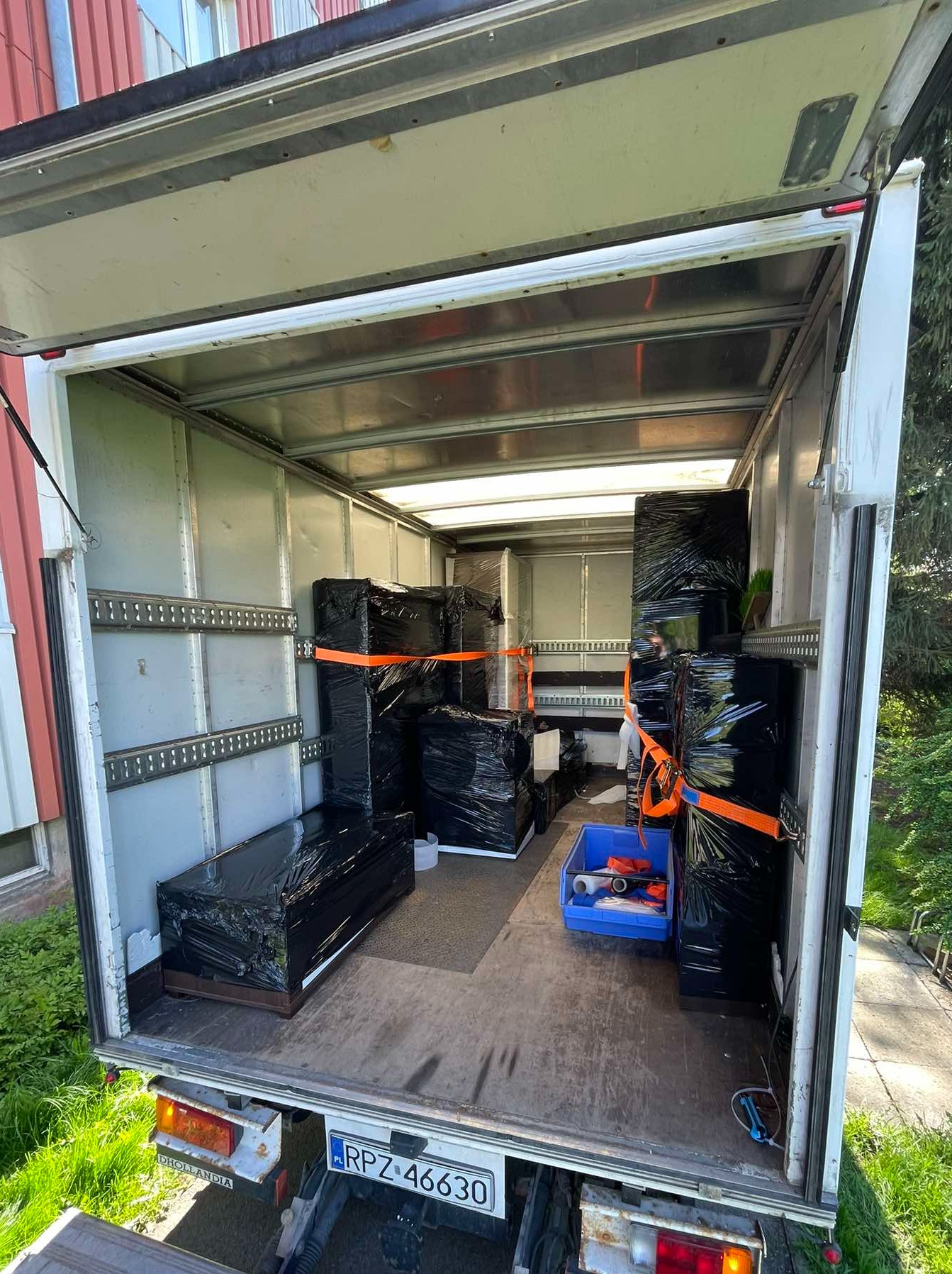 Wnętrze busa transportowego wypełnione meblami owiniętymi czarną folią i zabezpieczonymi pomarańczowymi pasami. Widoczny numer rejestracyjny pojazdu.