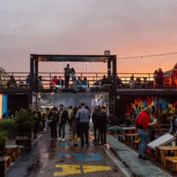 Plenerowa impreza z dwupoziomowymi konstrukcjami kontenerowymi, oświetloną figurą goryla i tłumem ludzi w wieczornym świetle.