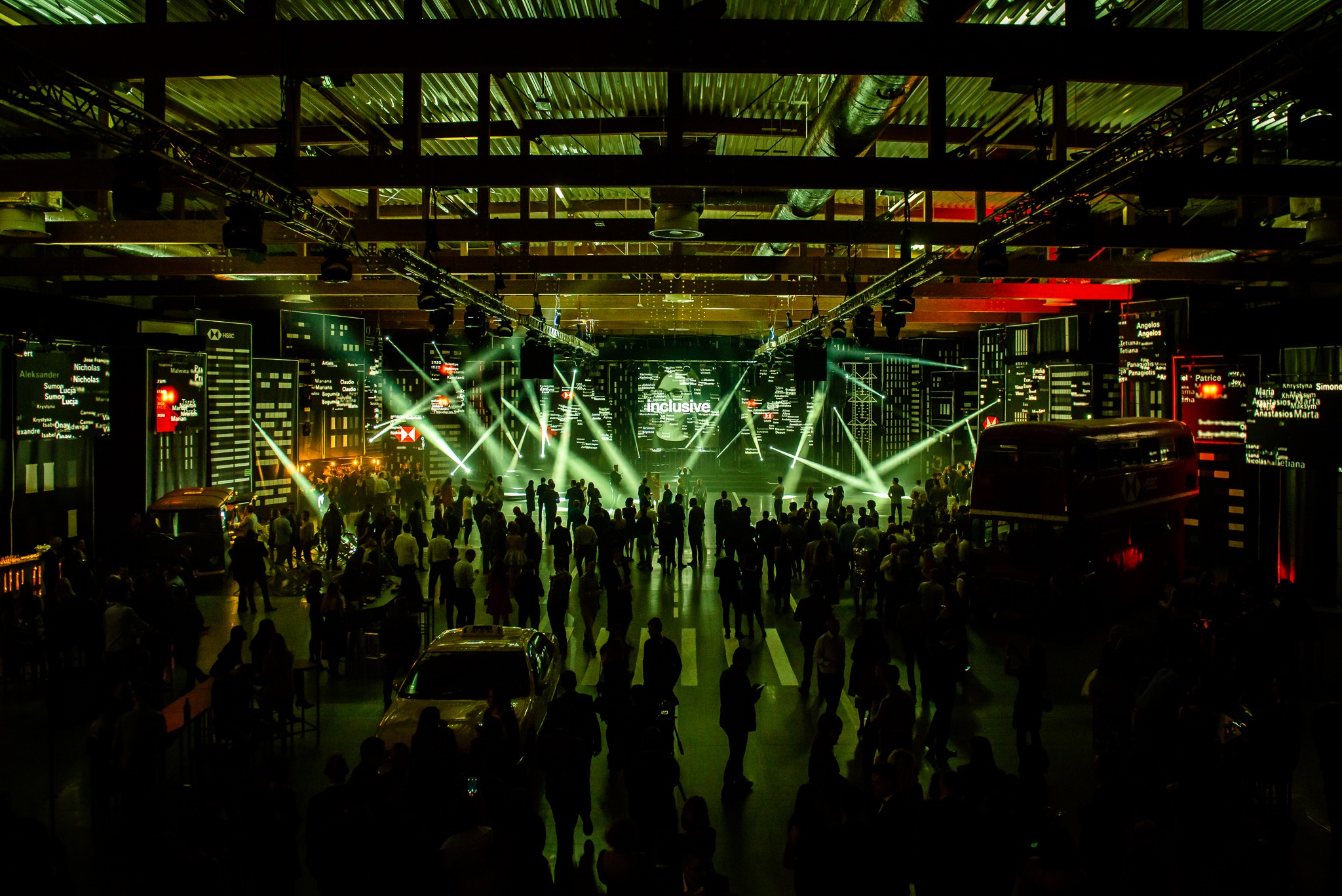 Widok z góry na dużą grupę ludzi w ciemnym pomieszczeniu z dekoracjami imitującymi miasto, oświetlonymi reflektorami i neonami, sugerujący imprezę firmową lub konferencję.