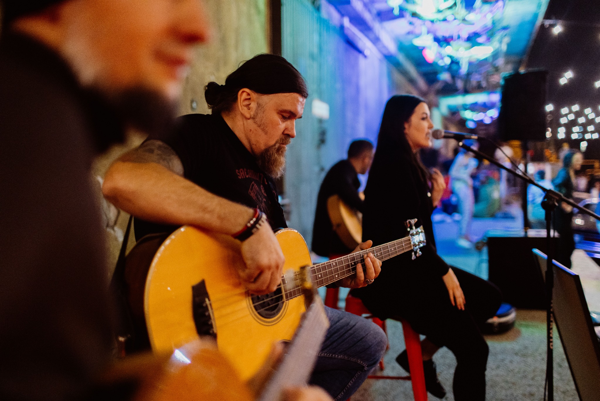 Zespół muzyczny w trakcie występu: gitarzysta gra na akustycznej gitarze, wokalistka śpiewa do mikrofonu na tle oświetlonej sceny i publiczności.