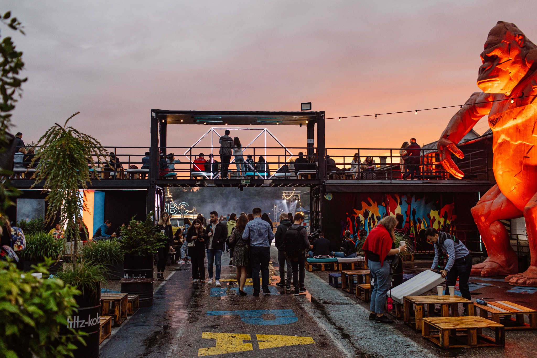 Plenerowa impreza z dwupoziomowymi konstrukcjami kontenerowymi, oświetloną figurą goryla i tłumem ludzi w wieczornym świetle.