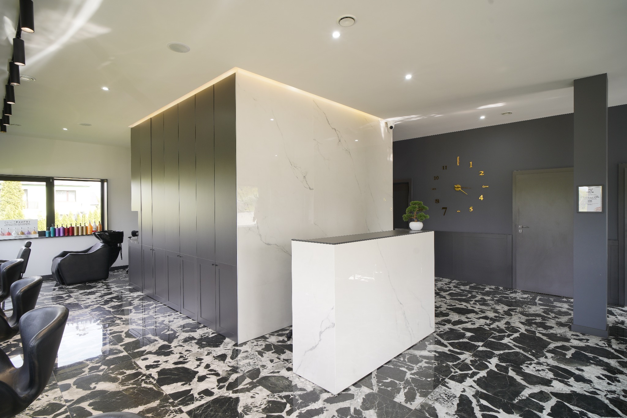 Nowoczesne wnętrze salonu fryzjerskiego z marmurową podłogą, białą recepcją, szarymi szafkami i ścianą z zegarem.