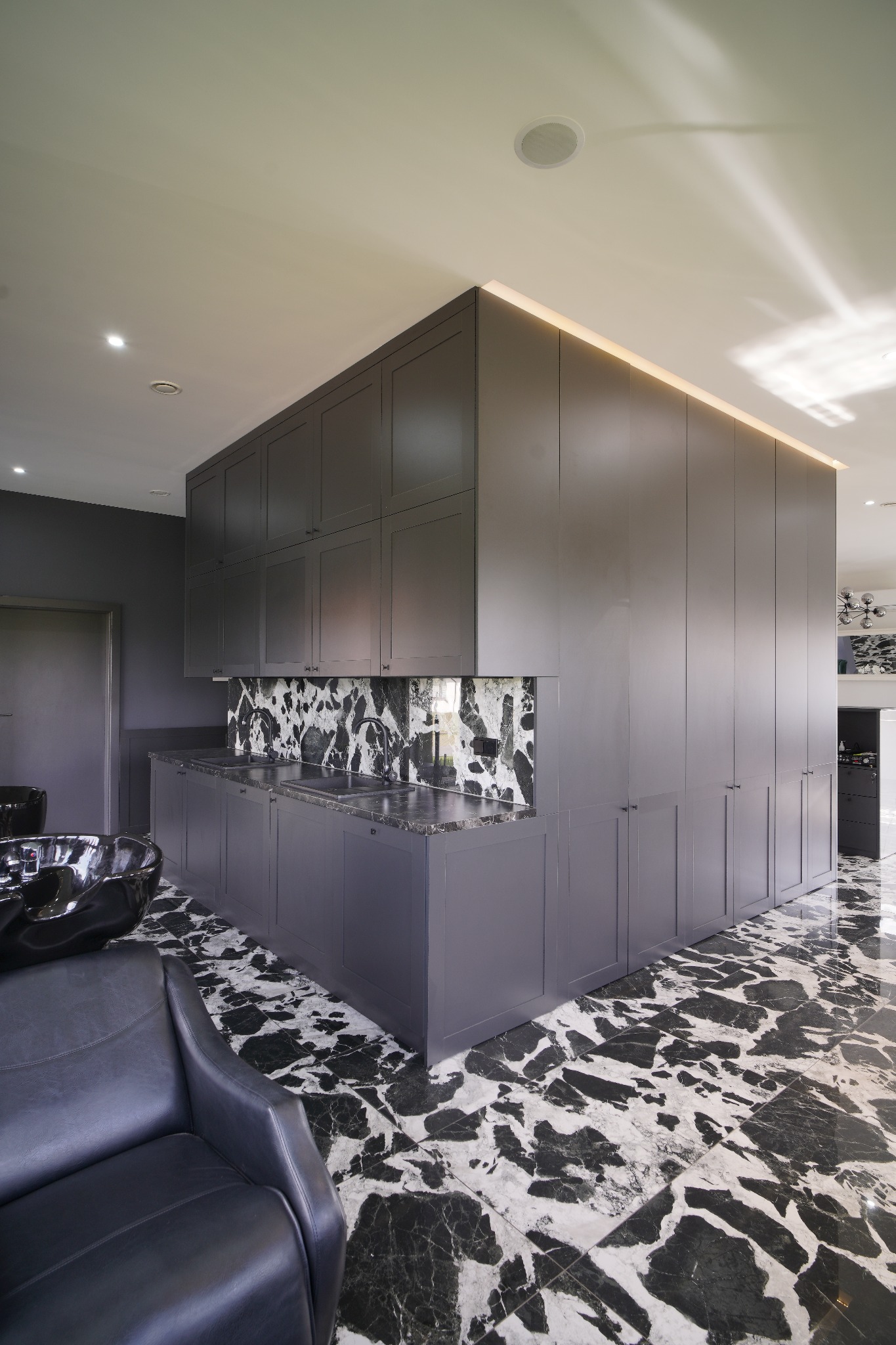 Wnętrze salonu fryzjerskiego z zabudową meblową w szarym kolorze, czarnym marmurem na podłodze i ścianie nad umywalkami, oraz czarnym fotelem fryzjerskim.