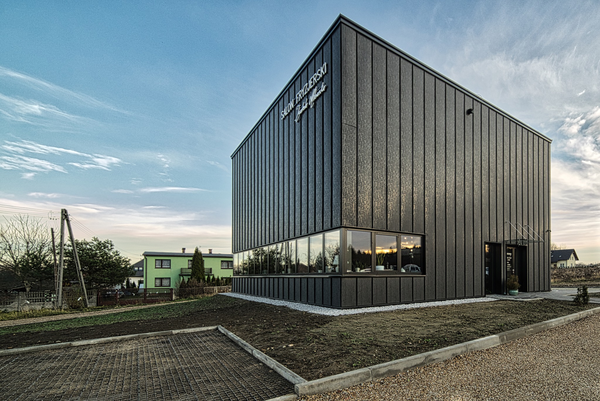 Nowoczesny, dwukondygnacyjny budynek usługowy z ciemną, pionowo żłobkowaną elewacją, oświetlony wieczornym światłem, z widocznym szyldem 'Salon Fryzjerski' nad oknami parteru.