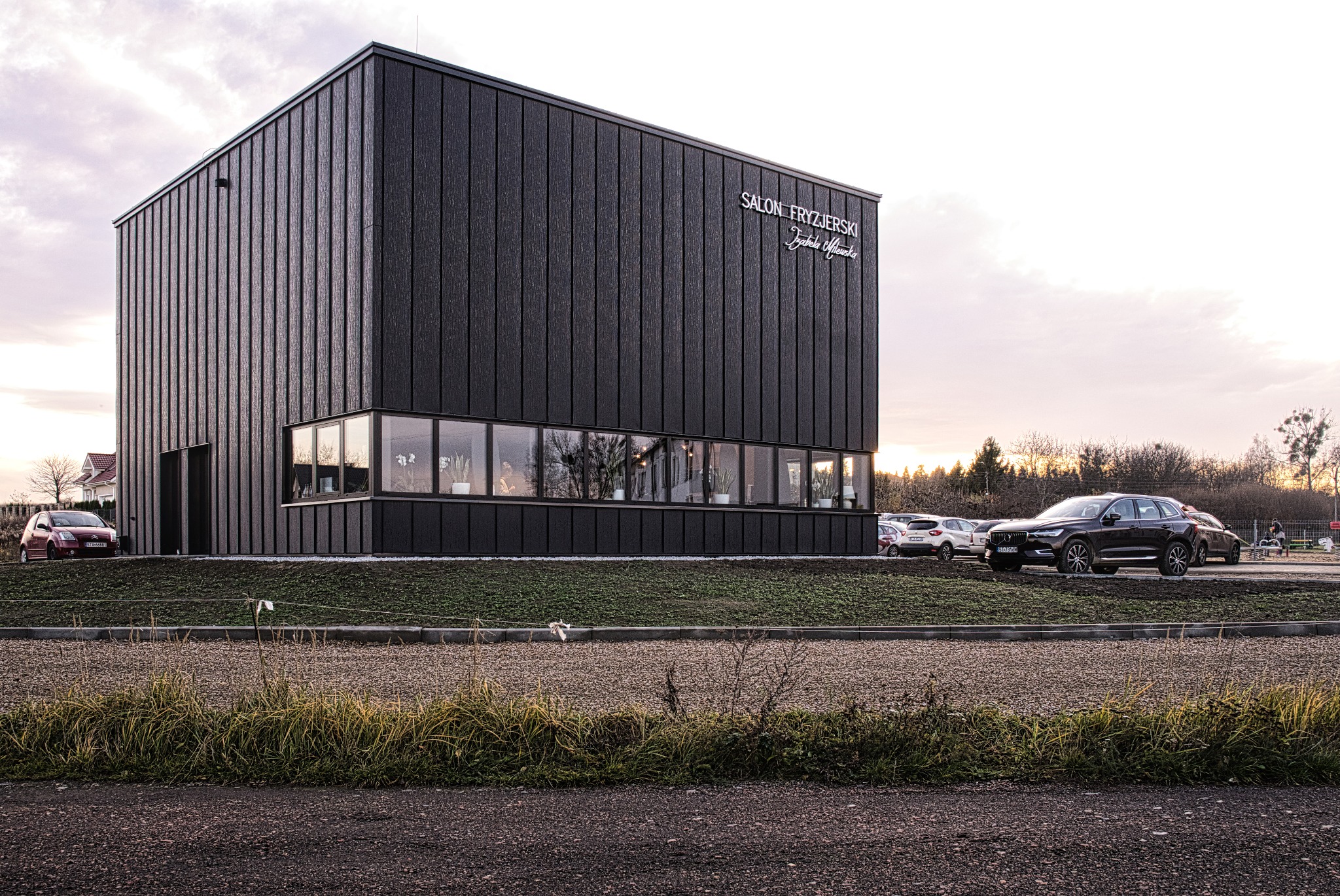 Nowoczesny budynek salonu fryzjerskiego z ciemną elewacją, dużymi oknami i napisem 'Salon Fryzjerski Izabela Olewska', otoczony parkingiem i zielenią, sfotografowany o zmierzchu.