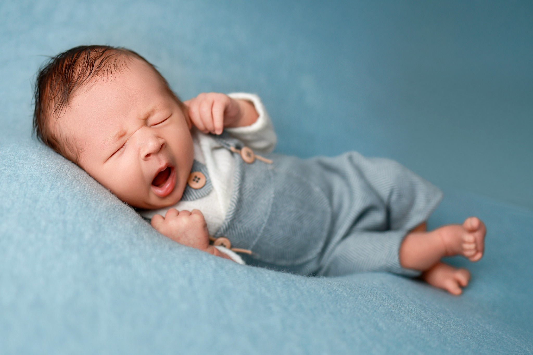 Noworodek w błękitnym ubranku śpi na miękkim, błękitnym tle, z otwartymi ustami i zaciśniętymi piąstkami.