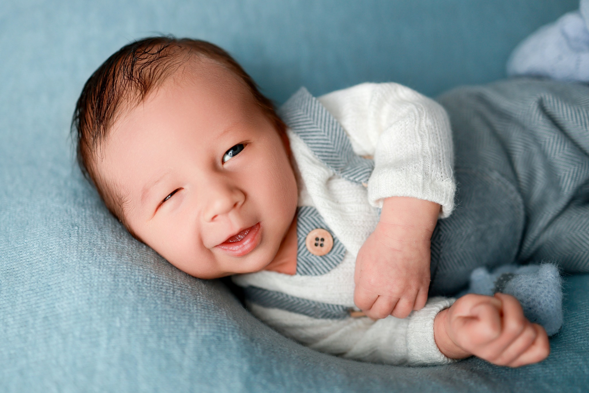 Uśmiechnięty noworodek w białym sweterku z drewnianym guzikiem i szarym kombinezonie, leżący na błękitnym kocyku, mruży jedno oko.