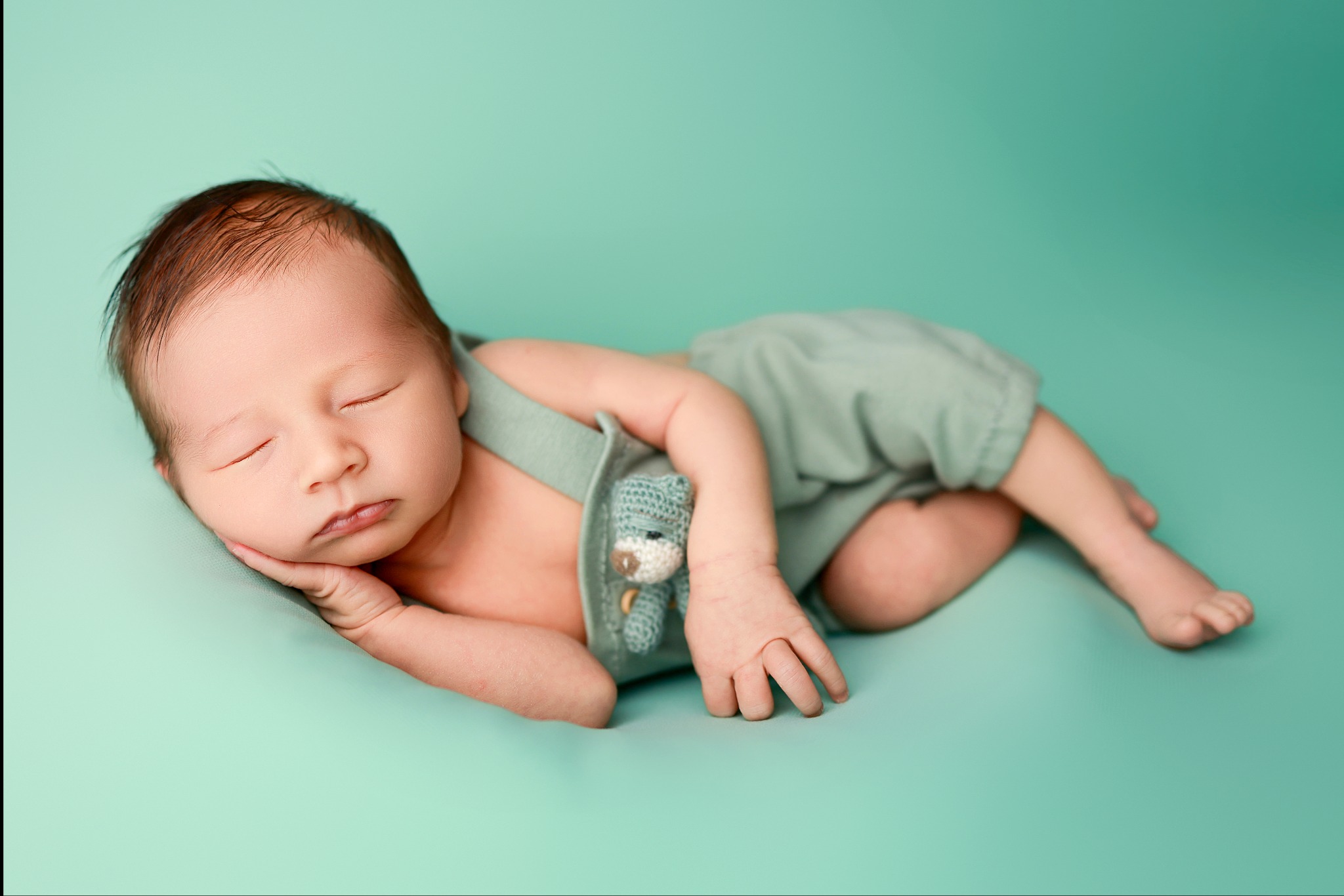 Śpiący noworodek w pastelowych, zielonych spodenkach i z dzierganym misiem, leżący na gładkim, turkusowym tle.