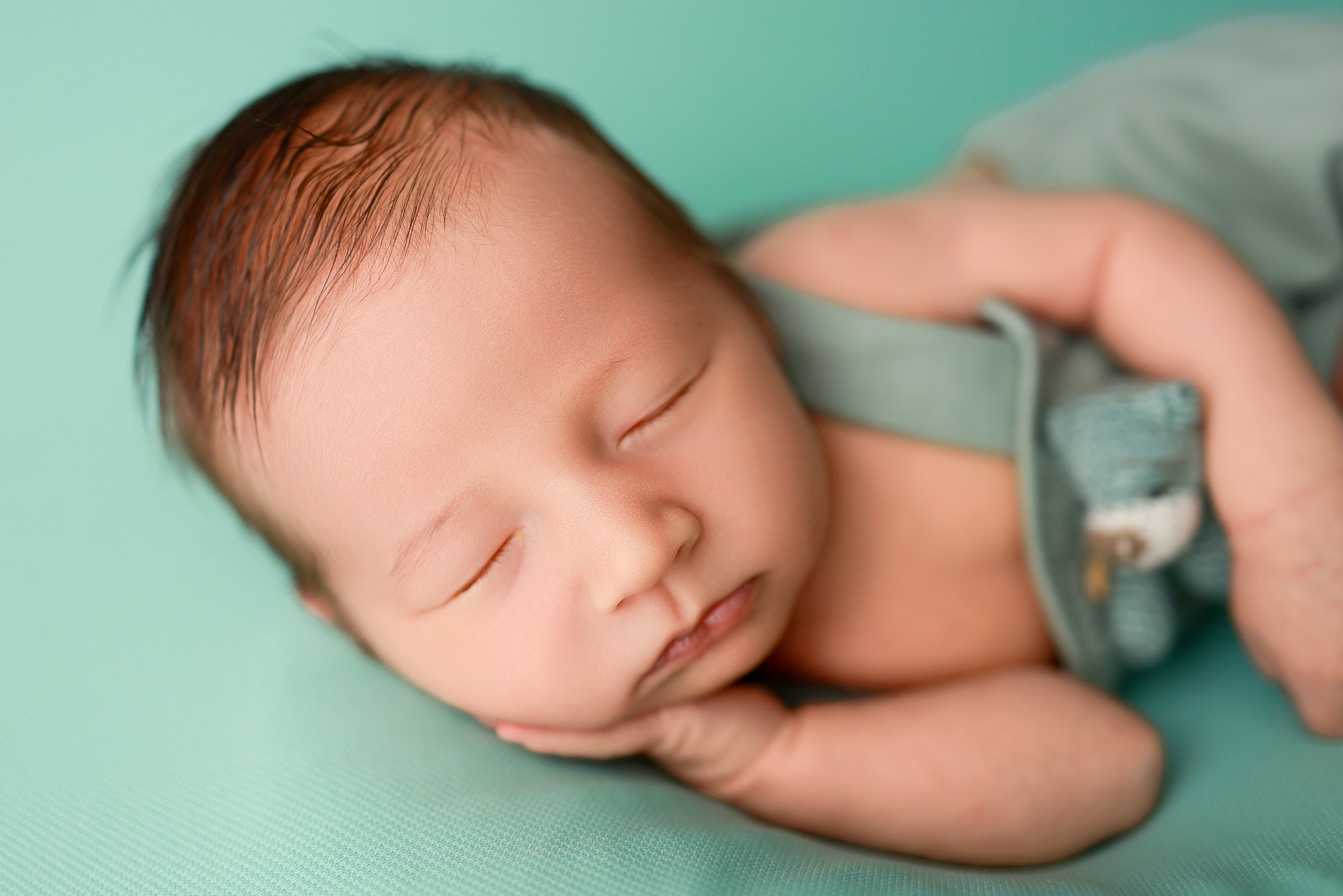 Śpiący noworodek w turkusowym ubranku z pluszowym misiem na turkusowym tle.