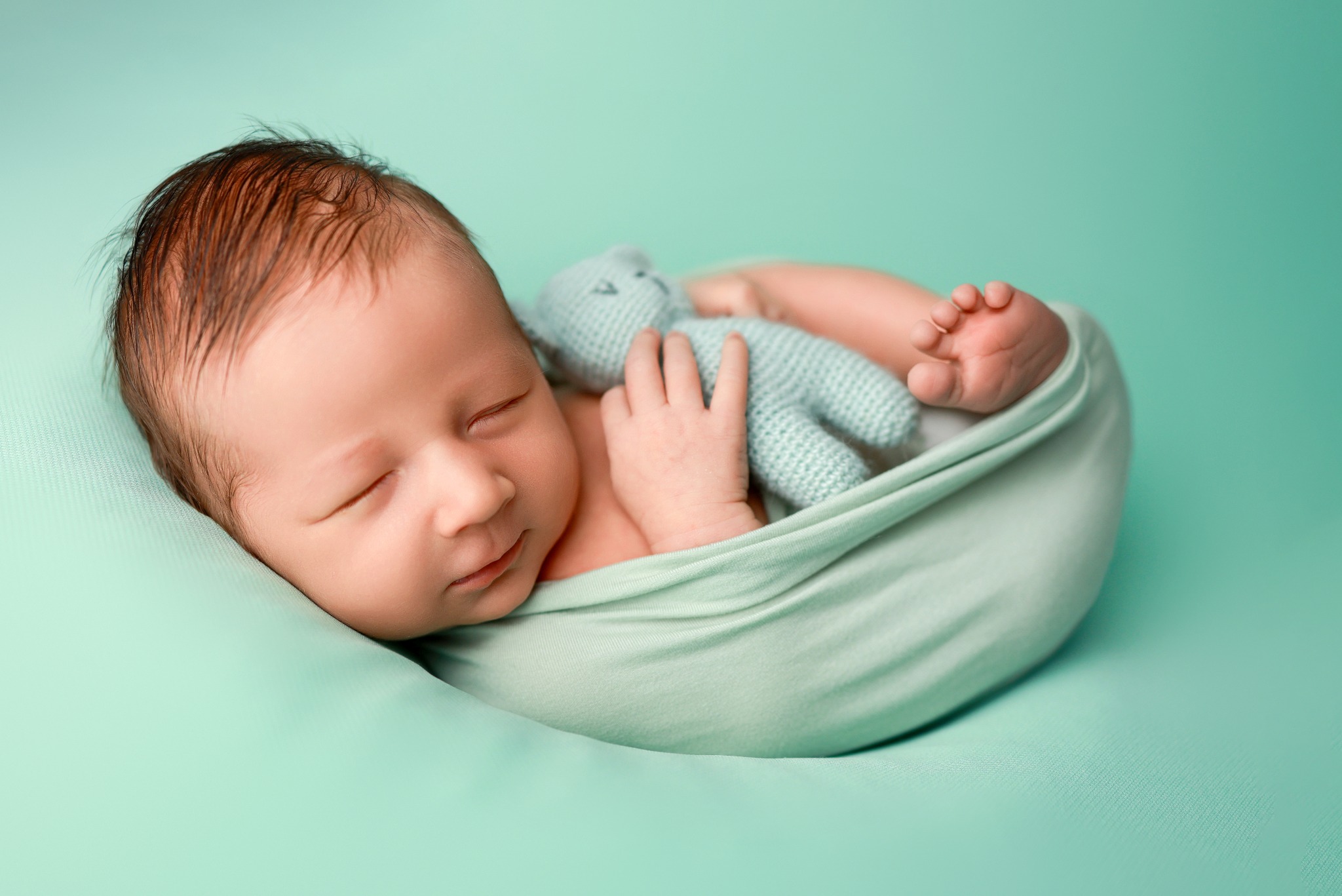 Noworodek śpi owinięty w miętowy materiał, trzymając w rączkach pluszowego, dzierganego misia w tym samym kolorze.