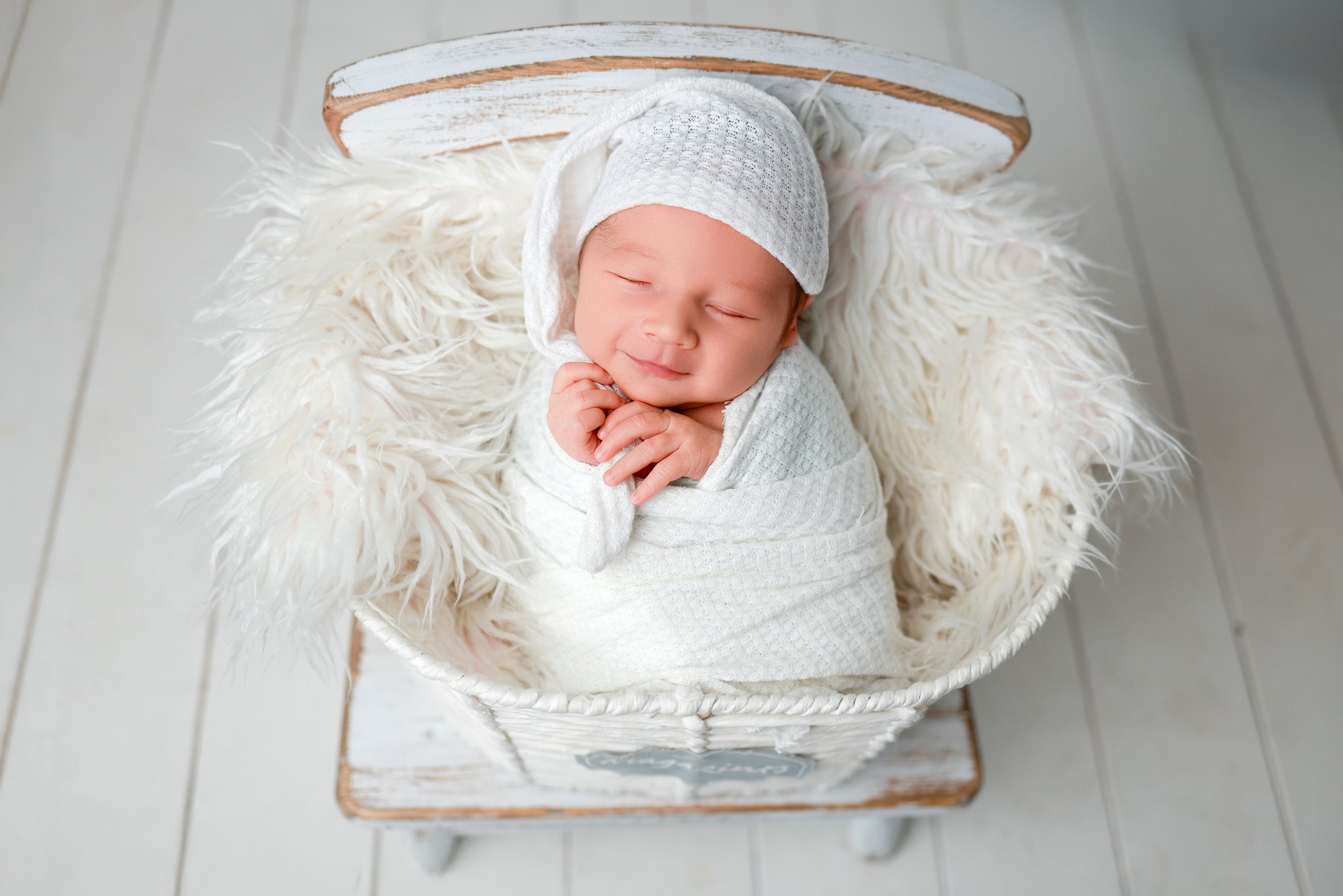 Śpiący noworodek w białym czapeczce i otulony w białym kocyku, leżący w białym, wiklinowym koszyku wypełnionym sztucznym futerkiem na tle białych desek.