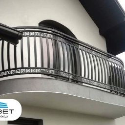 OWBET Bramy i Ogrodzenia - Czarna, metalowa balustrada balkonowa z motywem greckim na tle białej elewacji budynku. Widoczne logo firmy OW-BET w dolnej części kadru.