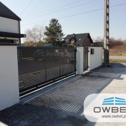 OWBET Bramy i Ogrodzenia - Nowoczesna, przesuwna brama wjazdowa z perforowanej blachy, w kolorze grafitowym, zamontowana przy białym murze posesji z numerem 20, w tle widoczny dom z ciemnym dachem.