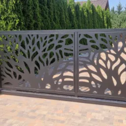 Czarna, przesuwna brama wjazdowa z ażurowym wzorem drzewa, zamontowana na posesji z kostki brukowej, obok skrzynka na listy z numerem domu 10A.