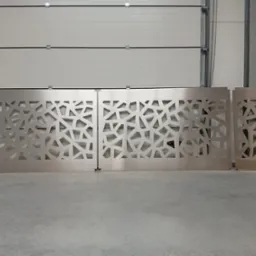 Metalowe panele z ażurowym wzorem geometrycznym, prawdopodobnie elementy dekoracyjne bramy garażowej lub wjazdowej, ustawione w hali.