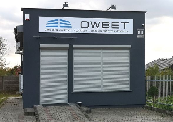 Fasada budynku firmy OWBET z szarymi roletami zewnętrznymi, oferującej akcesoria do bram i ogrodzeń, sprzedaż hurtową i detaliczną, widoczny numer budynku 84 przy ulicy Sobótki.