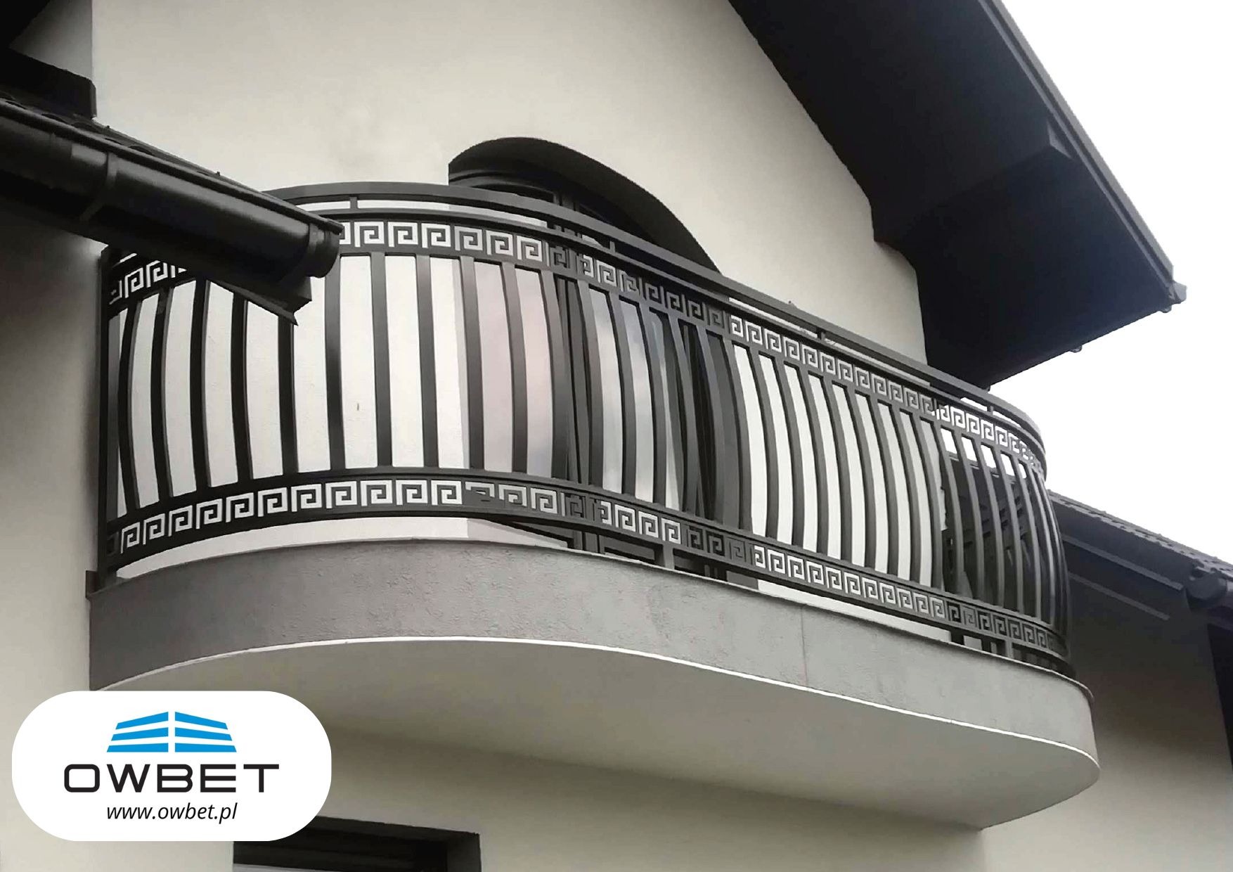 Czarna, metalowa balustrada balkonowa z motywem greckim na tle białej elewacji budynku. Widoczne logo firmy OW-BET w dolnej części kadru.