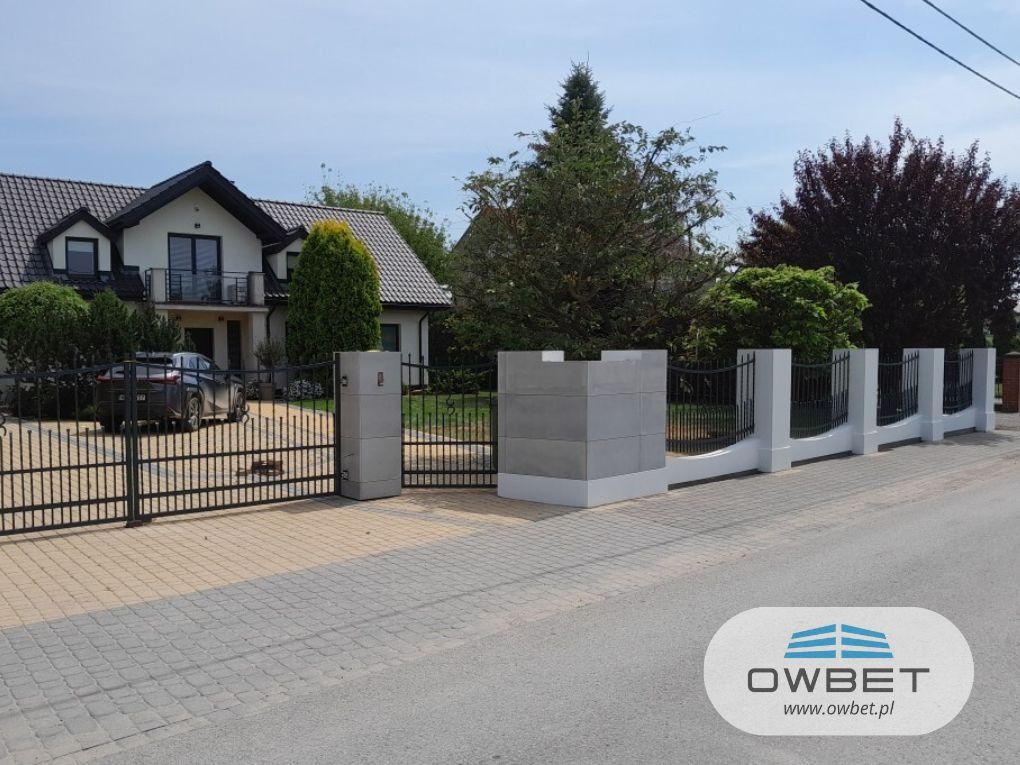 Nowoczesne ogrodzenie posesji z czarnymi, kutymi przęsłami i betonowymi słupkami, brama wjazdowa z automatyką, dom jednorodzinny w tle, widoczne logo producenta w prawym dolnym rogu.