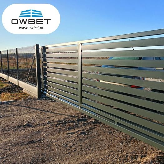 Nowoczesna przesuwna brama w kolorze szarym z poziomymi panelami, widoczna z boku, z fragmentem siatki ogrodzeniowej w tle i samochodem za bramą.