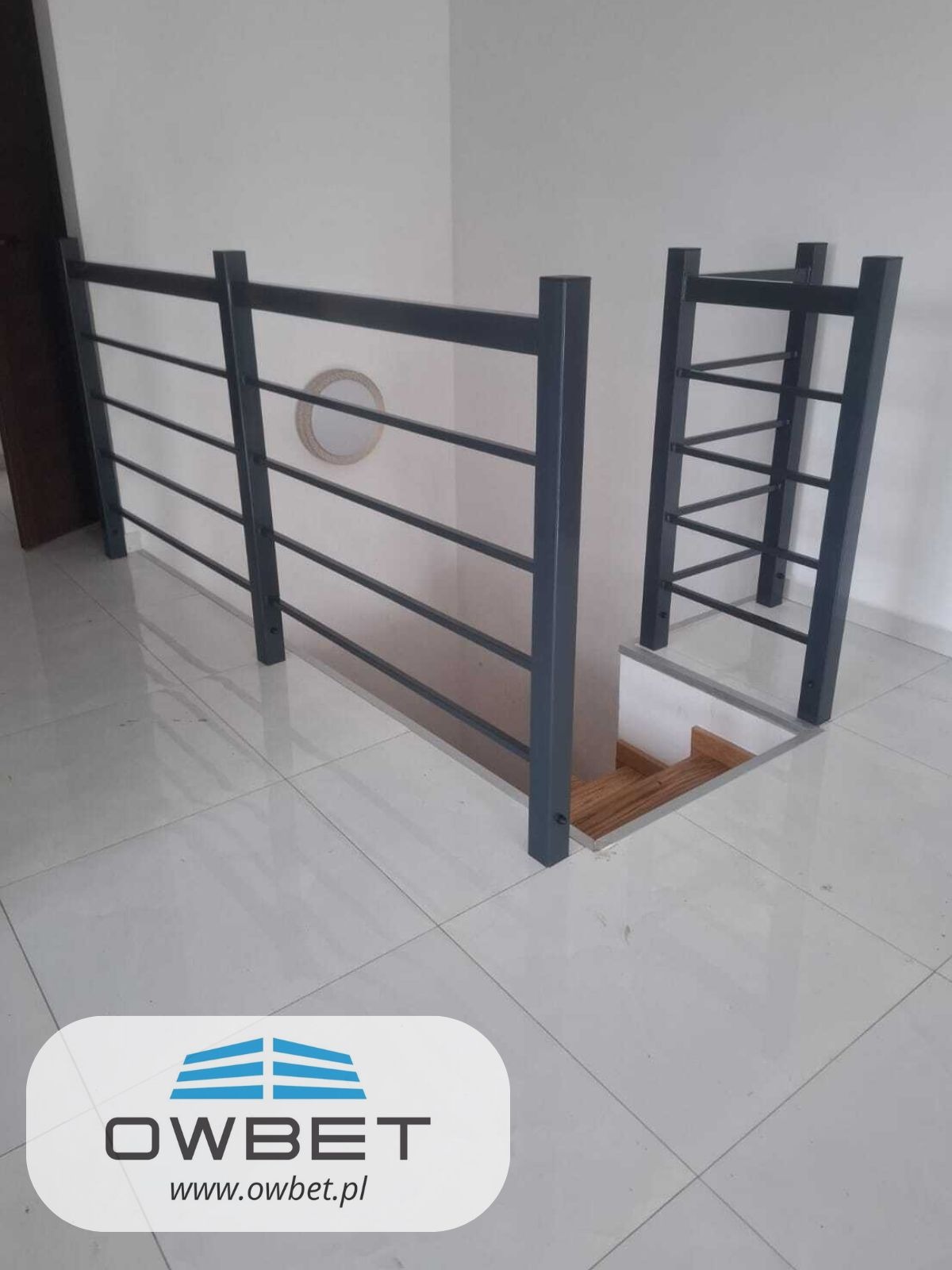 Szara, metalowa balustrada schodowa z poziomymi prętami i drabinką na antresoli, widok z góry na białe płytki podłogowe.