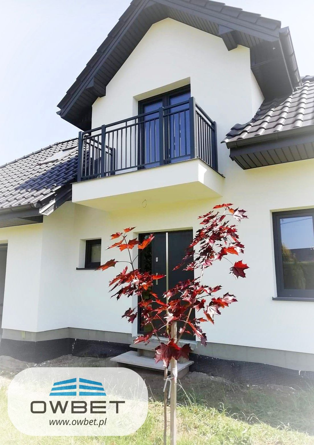 Biały dom z czarną stolarką okienną i balkonem z metalową balustradą, na pierwszym planie młody klon palmowy o czerwonych liściach, widoczne logo firmy w rogu.