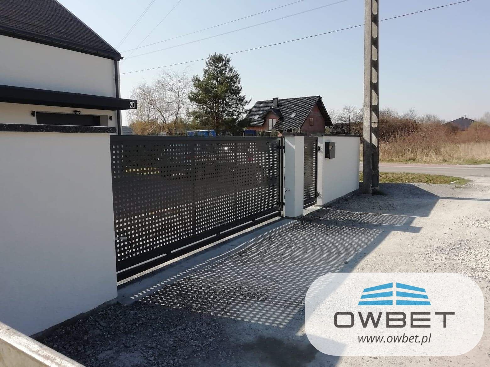 Nowoczesna, przesuwna brama wjazdowa z perforowanej blachy, w kolorze grafitowym, zamontowana przy białym murze posesji z numerem 20, w tle widoczny dom z ciemnym dachem.