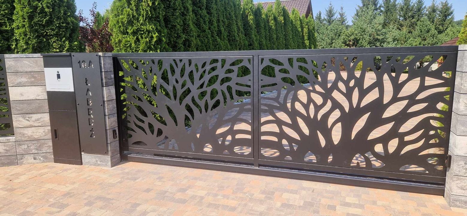 Czarna, przesuwna brama wjazdowa z ażurowym wzorem drzewa, zamontowana na posesji z kostki brukowej, obok skrzynka na listy z numerem domu 10A.