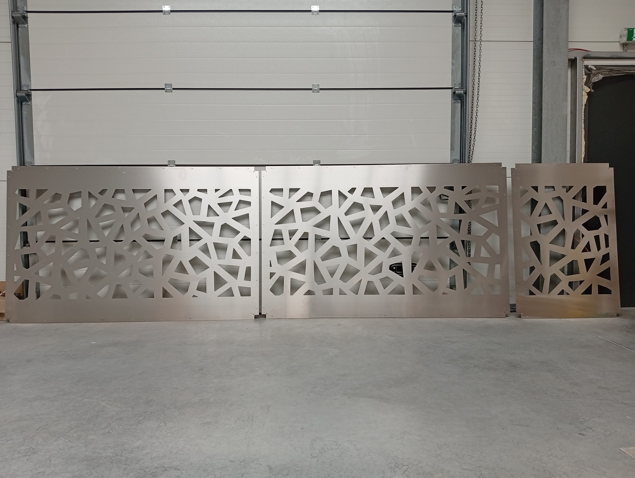 Metalowe panele z ażurowym wzorem geometrycznym, prawdopodobnie elementy dekoracyjne bramy garażowej lub wjazdowej, ustawione w hali.