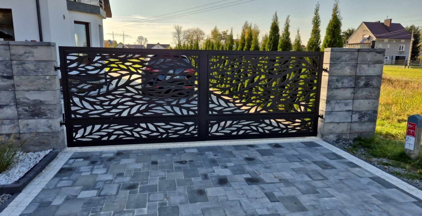 Nowoczesna brama wjazdowa z ażurowym wzorem liści na posesji z kostką brukową i słupkami z szarego kamienia. W tle dom i zieleń.