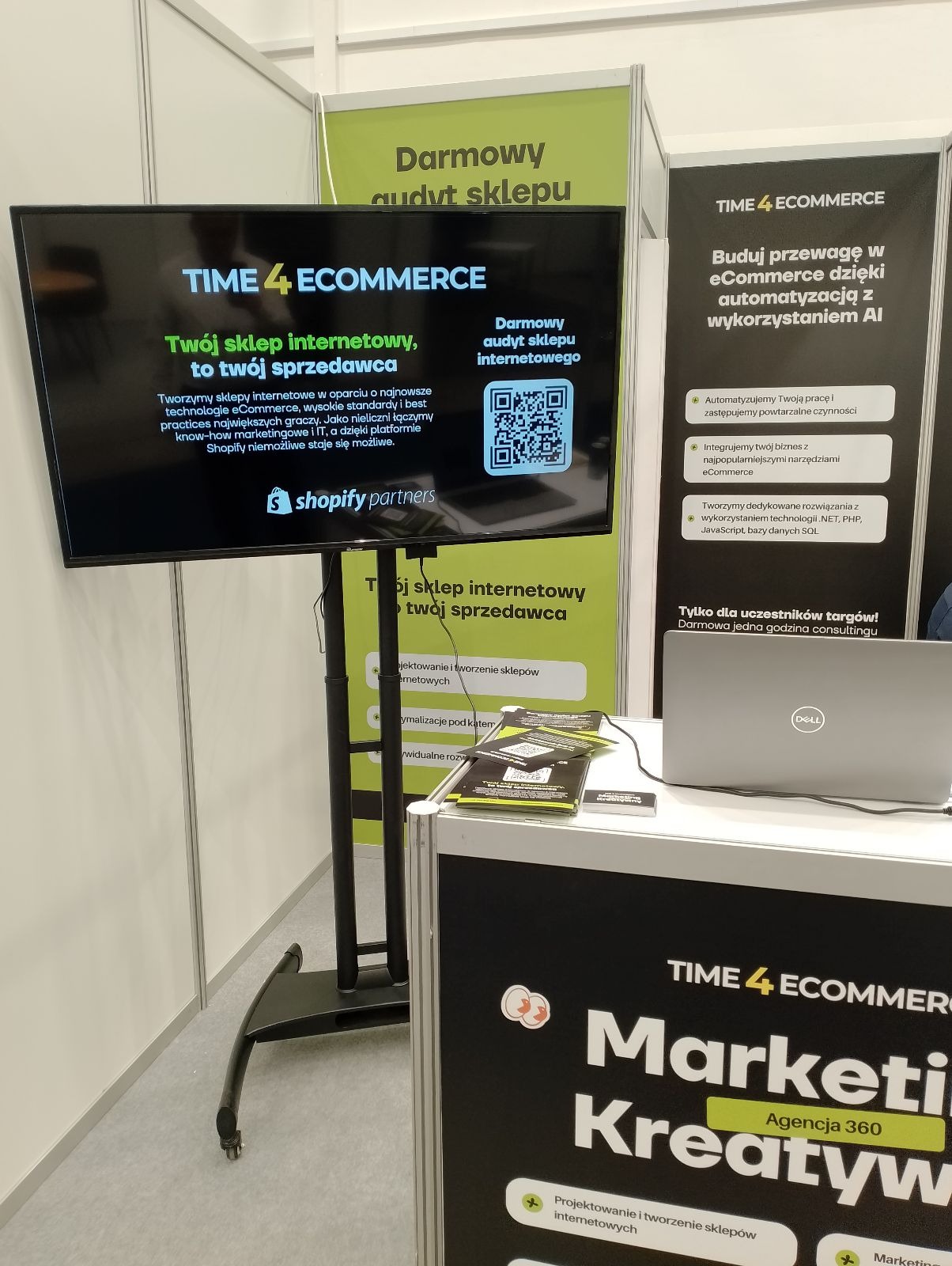 Ekspozycja targowa firmy Time4Ecommerce z ekranem prezentującym ofertę tworzenia sklepów internetowych i kod QR do darmowego audytu, obok baner z hasłem o przewadze w eCommerce dzięki AI i laptop...