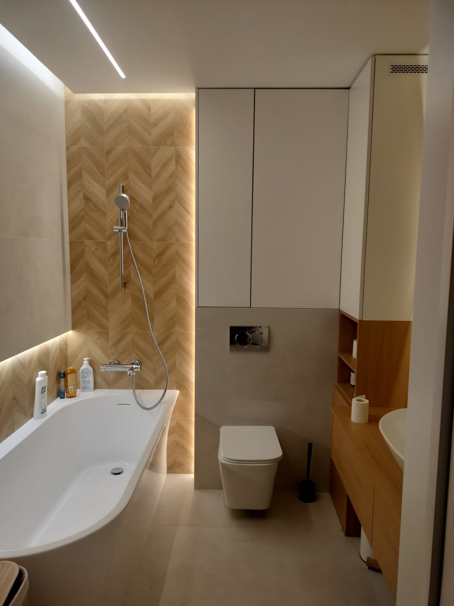 Nowoczesna łazienka z wanną, toaletą podwieszaną i drewnianą zabudową umywalki, podświetlona ledami, z akcentem na ścianę z płytek ułożonych w jodełkę.