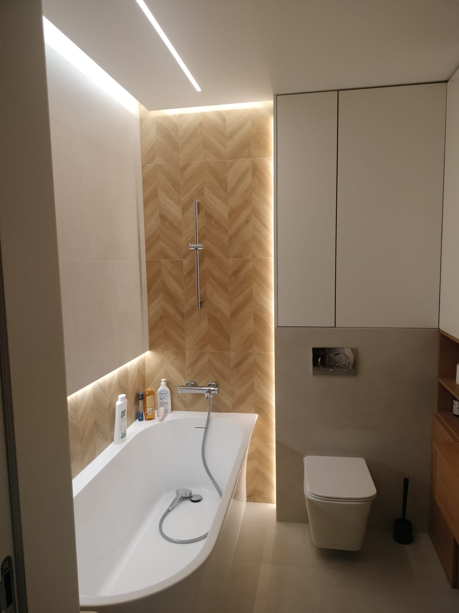 Nowoczesna łazienka z podświetlaną ścianą w jodełkę, białą wanną z prysznicem ręcznym, podwieszaną toaletą i szafkami w zabudowie.